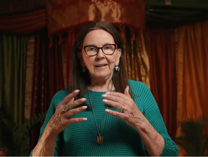 Elderly woman speaking in colorful draped room.